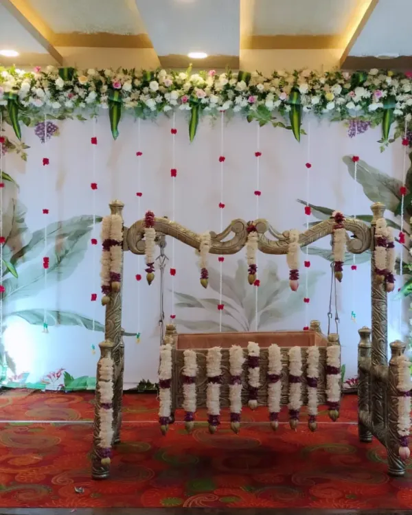 A traditional naming ceremony setup featuring a beautiful floral frame around a decorated cradle. The backdrop uses soft fabrics and hanging flowers to create a serene and sacred atmosphere.