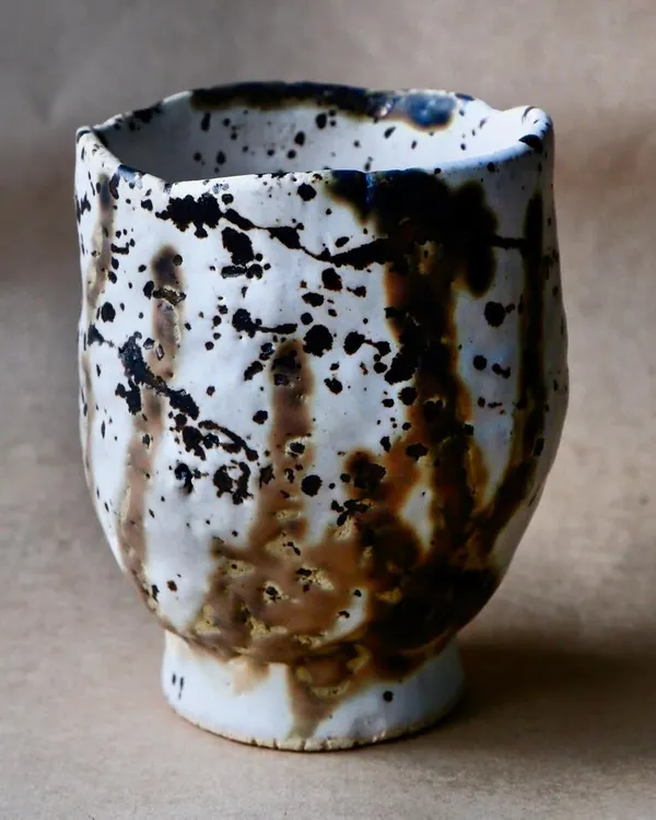 This handmade ceramic cup features a dynamic splash of black and brown glaze over a white base. The irregular rim adds to its unique, handcrafted character.