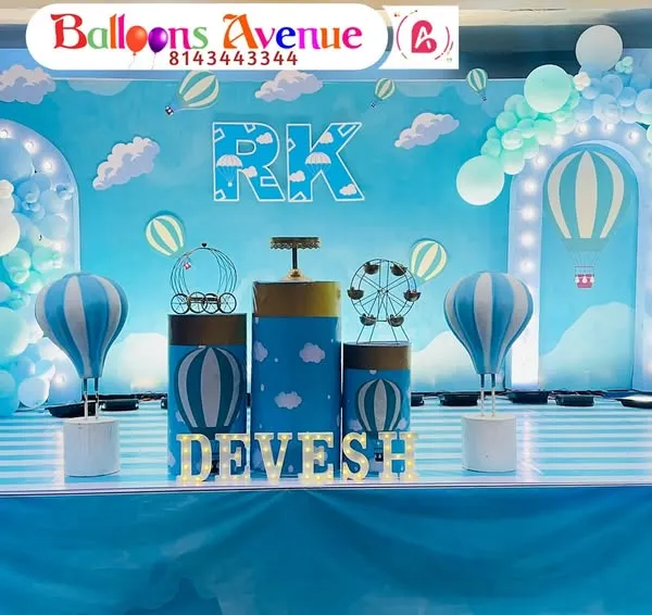 A charming hot air balloon theme for Devesh's birthday. The stage is decorated with a sky-blue backdrop, fluffy cloud cutouts, and a striped floor to complete the look.