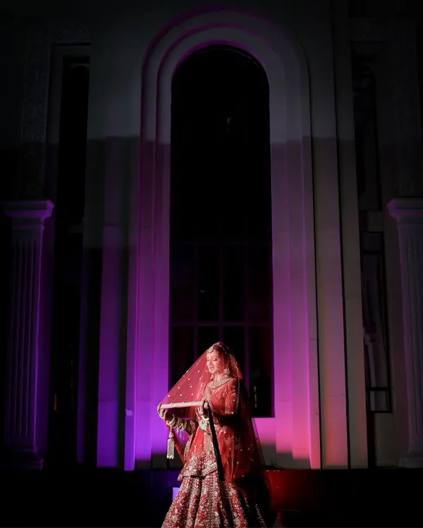 A dramatic bridal portrait using purple uplighting and architectural elements. The bride, in her red lehenga, looks regal and mysterious, like a queen in her palace.
