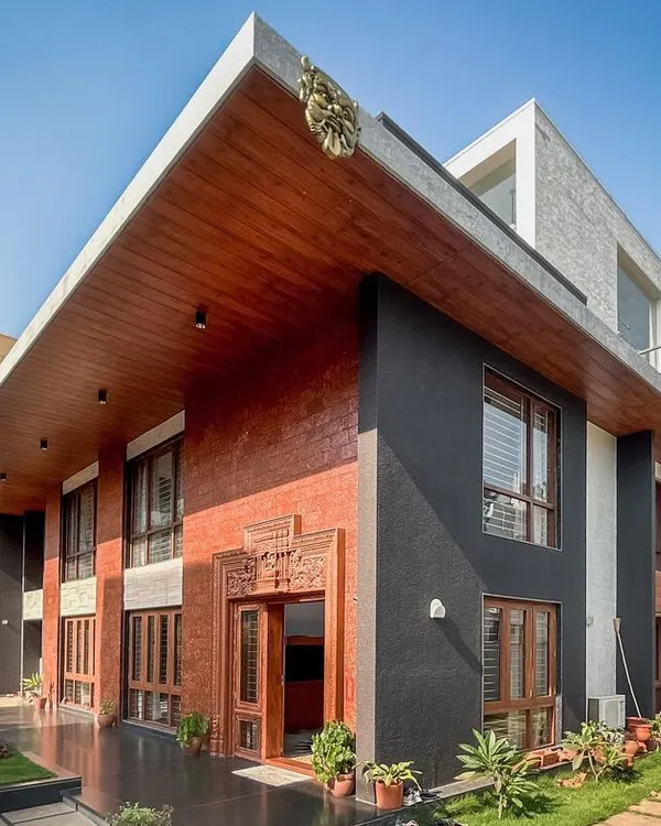 The facade of a contemporary villa in Hennur, showcasing a dynamic composition of exposed brick, dark textured surfaces, and a prominent cantilevered roof.