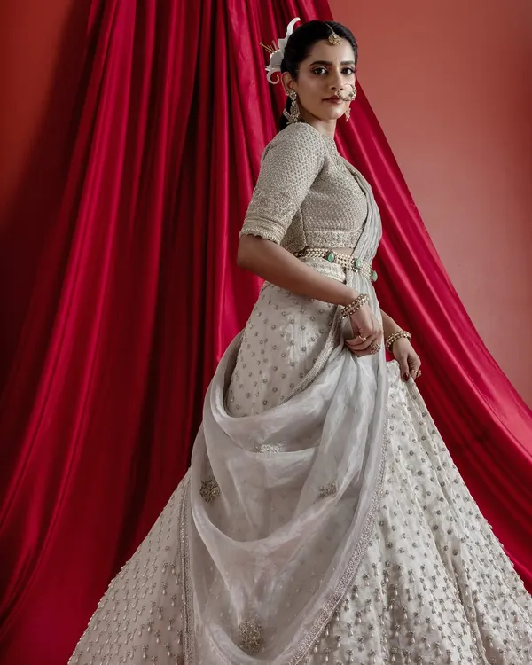 This ethereal ivory lehenga is the embodiment of modern bridal elegance. The voluminous skirt is covered in intricate embroidery and delicate pearl highlights, paired with a meticulously hand-worked blouse and a statement emerald and pearl belt, creating a look of timeless grandeur.