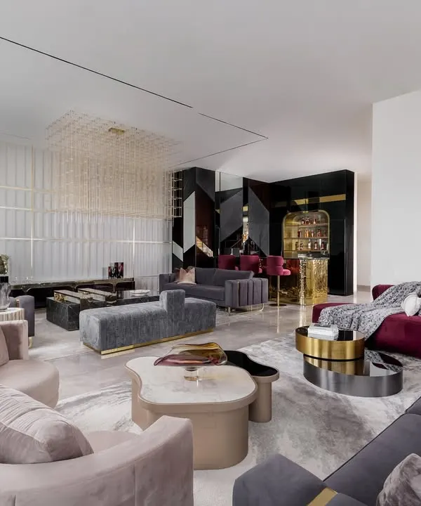 An expansive view of a complete entertainment lounge we designed for hosting and relaxation. This space features multiple seating zones with custom curved and channel-tufted sofas, a sculptural grid chandelier, and a dedicated bar area, all tied together with a palette of greys, blush, and rich burgundy.