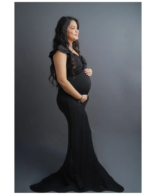 A timeless and elegant solo maternity portrait. This side profile shot beautifully showcases the baby bump, with the mother-to-be in a classic black gown against a solid grey studio background.