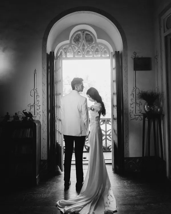 A dramatic, cinematic portrait of a couple standing in a grand doorway. The high contrast black and white emphasizes their elegant silhouettes and the promise of the moment.