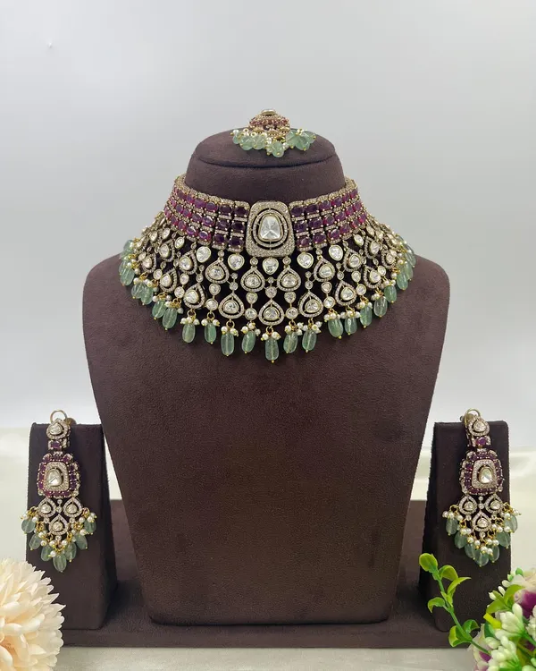 The complete view of a premium bridal choker set. It features rows of ruby-colored stones, a layer of uncut polki, and mint-green bead drops, combining classic colors with a grand design. The set includes the choker, earrings, and maang tikka.