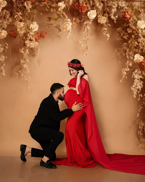 A classic and romantic maternity pose. The partner's gentle kiss on the baby bump against a floral-draped, fine art backdrop creates a timeless and passionate portrait. The vibrant red gown adds a touch of glamour to this special moment.