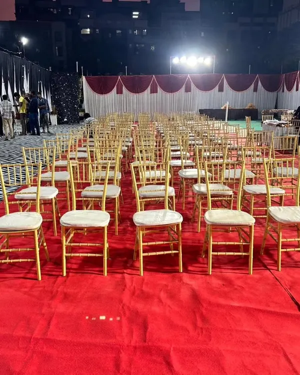 An outdoor wedding reception setup showing the guest seating arrangement. We used elegant gold chiavari chairs neatly arranged on a red carpet, with draped tents in the background, ready for an evening celebration.