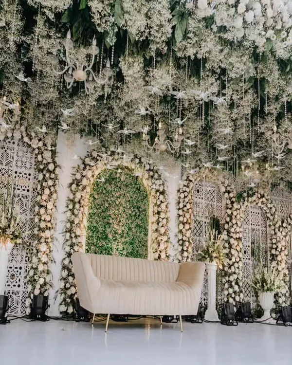 This all-white engagement stage is the definition of timeless elegance. I used a lush green backdrop as a canvas and layered it with thousands of white roses, baby's breath, and hanging floral installations to create a dreamy, ethereal atmosphere. The intricate jaali panels and a mirrored aisle add a touch of modern royalty.