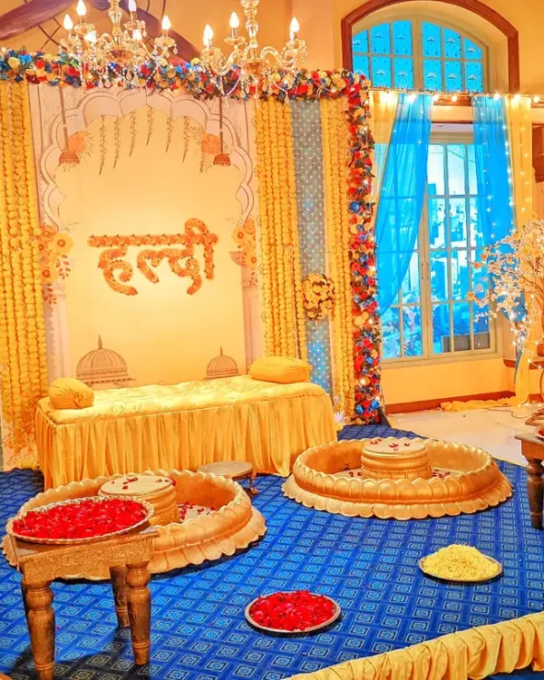 A grand Haldi ceremony setup with a royal theme, featuring golden statues, a decorated divan, and traditional brass urlis filled with flower petals.