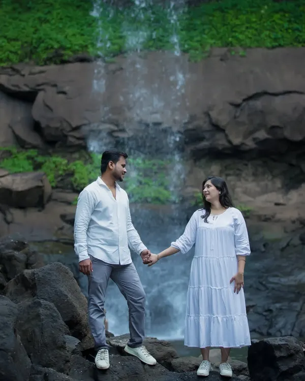 An expecting couple shares a look of love while holding hands, standing on rocks in front of a cascading waterfall. Dressed in coordinated white outfits, this maternity photo has a serene and epic feel, set against a stunning natural landscape.