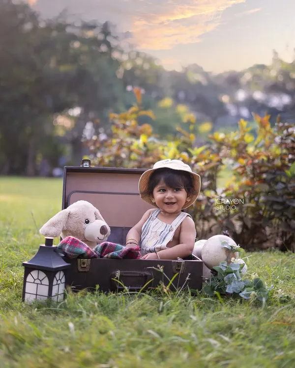 A little explorer is ready for an adventure. This baby boy smiles from inside a vintage suitcase, surrounded by his travel companions in a sunlit park.