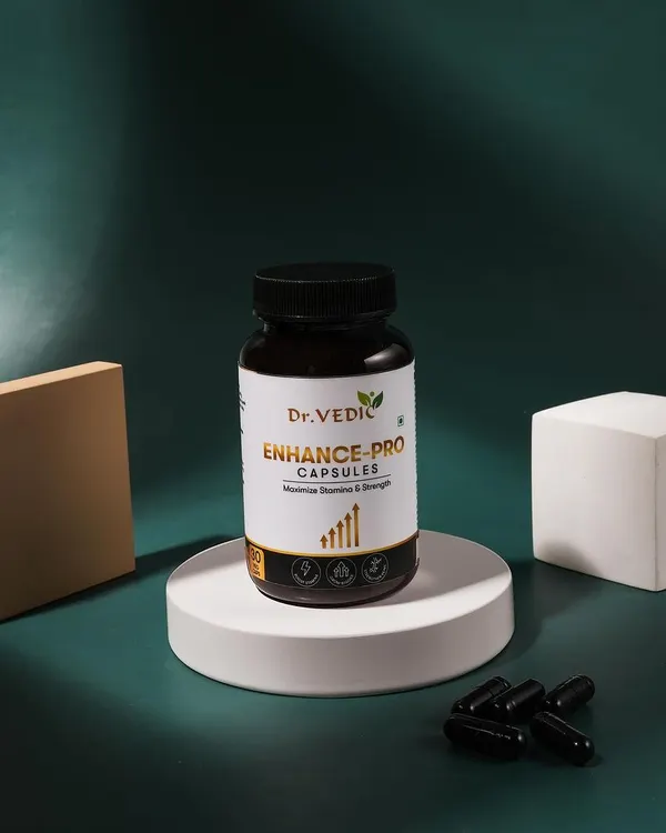 A clean and minimalist shot of Dr. Vedic Enhance-Pro capsules. The product is placed on a white podium against a dark green background, with dramatic lighting to create a premium feel.