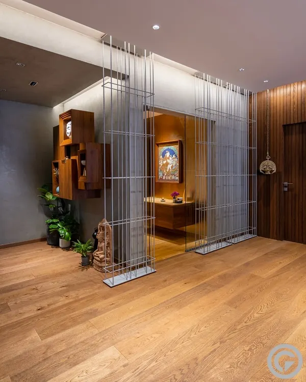 In this apartment foyer, a transparent screen of metal rods separates the entrance from the sacred puja room, creating a visual partition that maintains a sense of openness. The design layers modern materials with traditional art and warm wood flooring.
