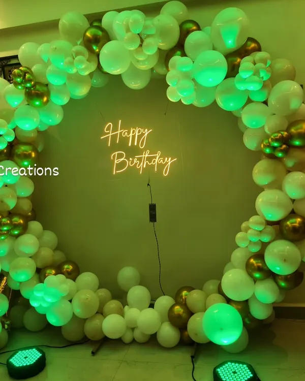 A classic and elegant ring backdrop for a birthday celebration. This design uses a full circle of white and chrome gold balloons, lit by a green spotlight, with a 'Happy Birthday' neon sign at the center for a modern and chic look.