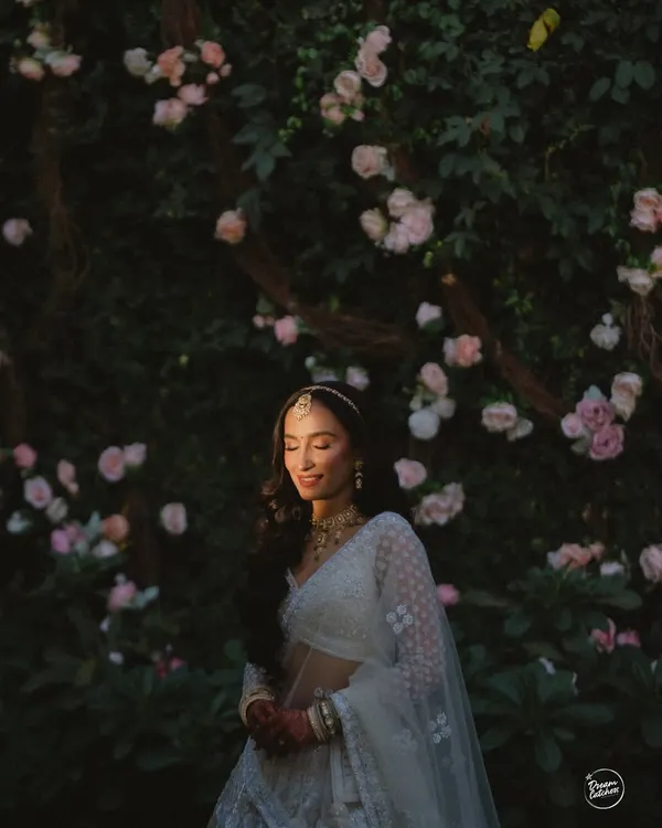 A portrait of a bride bathed in golden light, standing before a wall of blush pink roses. Our floral backdrops are designed to create these soft, romantic, and painterly moments.