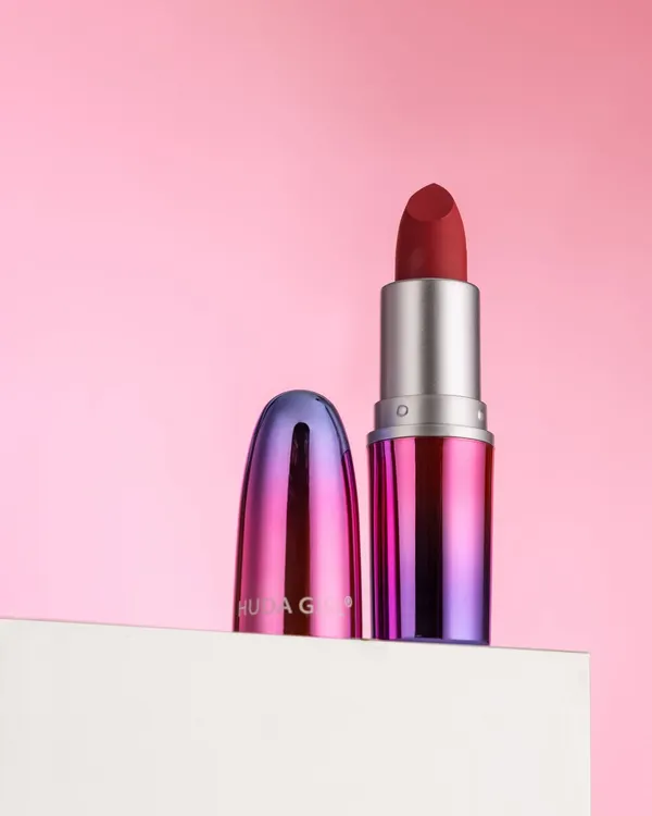 A clean and elegant shot of a Huda Girl lipstick against a soft pink background. The composition is simple, modern, and puts all the focus on the product's unique packaging.