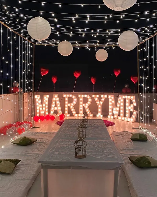 A romantic rooftop proposal setup featuring large "MARRY ME" marquee letters, low seating for an intimate dinner, and a canopy of warm fairy lights creating a magical atmosphere.