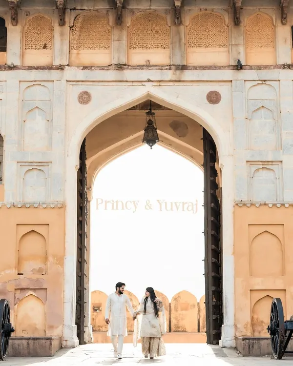 A grand entrance at Amer Fort in Jaipur. The couple, dressed in coordinated white traditional outfits, walks hand-in-hand through the magnificent archway, creating a timeless and regal portrait.