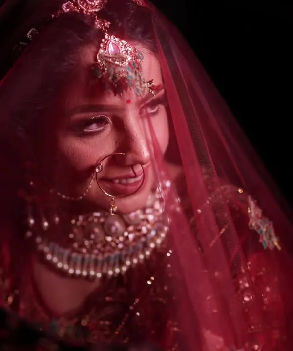 This is the style of bridal portrait I love to create. Using moody, dramatic lighting, I focus entirely on the bride, capturing that quiet moment of anticipation under the red veil. The rich colors of her lehenga and the intricate details of her jewelry, like the nath and maang tikka, stand out beautifully against the dark background.