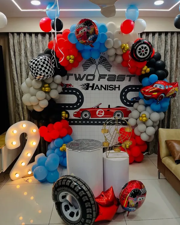 A "Two Fast" racing car theme for a second birthday. This setup includes a custom backdrop with a racetrack, a circular balloon arch in red, black, and white, and a large light-up number '2'.