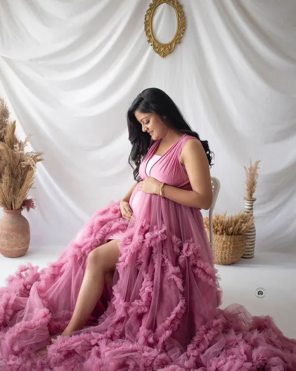 A quiet moment of reflection for this beautiful mama-to-be, seated in a stunning ruffled pink gown that fills the frame with color and texture.
