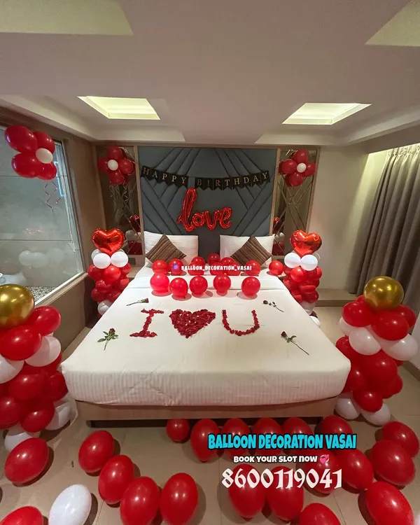 A classic romantic room decoration in red and white. The setup includes balloon pillars, a "love" foil balloon, and "I ❤️ U" written with rose petals on the bed.