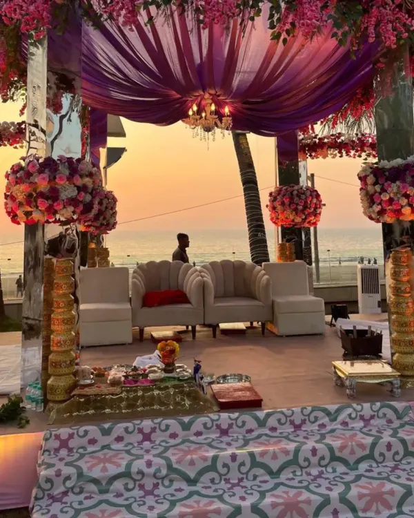 This is the kind of magic we create for a beachside engagement ceremony. The setup features a modern mandap with mirrored pillars, lush purple and pink floral arrangements, and elegant seating, all set against a breathtaking Mumbai sunset over the ocean.
