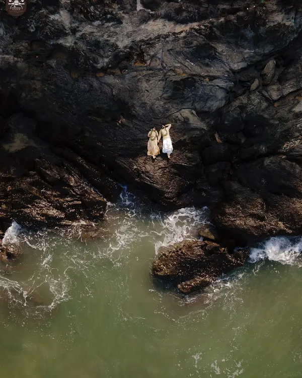 An aerial drone shot captures a couple standing on the edge of dramatic coastal rocks, with green waves washing ashore. This pre-wedding photograph highlights my ability to blend epic landscapes with intimate moments for a truly cinematic feel.