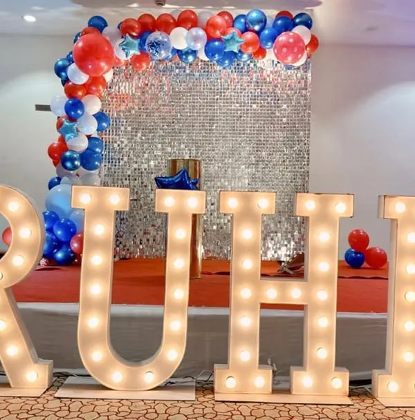 We provide large, illuminated marquee letters to personalize any event. Here, the name 'RUHI' is spelled out in lights, creating a bold and impressive focal point in front of a silver sequin wall and balloon arrangement.