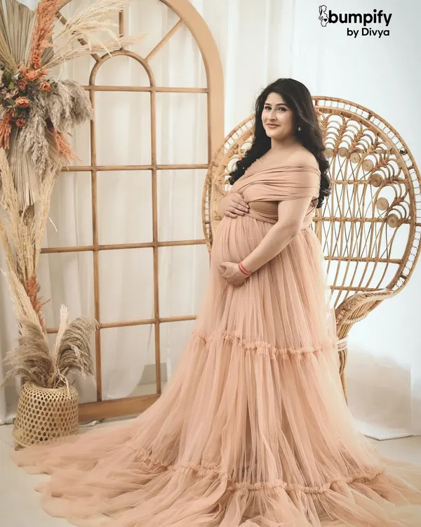 A serene solo portrait of an expecting mother in a beautiful peach gown, seated on a boho-style wicker chair. This shot is all about capturing that quiet, powerful glow of motherhood.