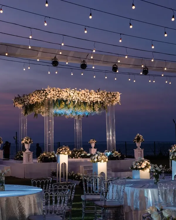 A fairytale sundowner mandap in Goa. This setup features a modern acrylic structure adorned with white florals, set against the purple and orange hues of the sunset, with strings of fairy lights creating a magical ambiance.