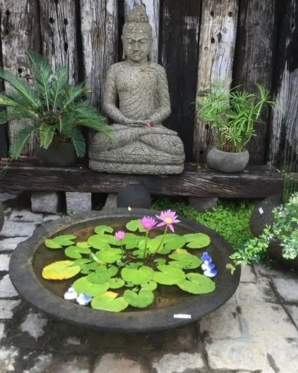 A tranquil garden scene featuring a stone Buddha statue and a floating lotus pond. The rustic wooden background and surrounding plants create a natural and calming environment.