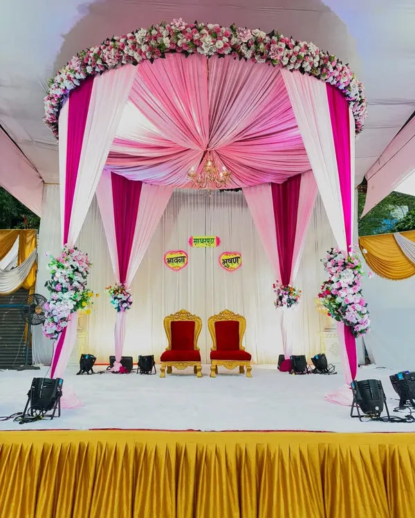A beautiful mandap setup perfect for an engagement ceremony. The pink and white drapes create a royal canopy, complemented by a small chandelier and lush floral arrangements, with traditional red chairs for the couple.