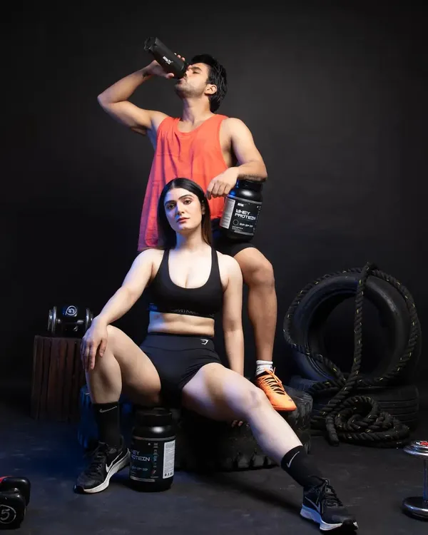 A full lifestyle scene for a fitness brand, featuring male and female models in a gym setting with whey protein products. This shot is designed to create an aspirational connection with the customer.