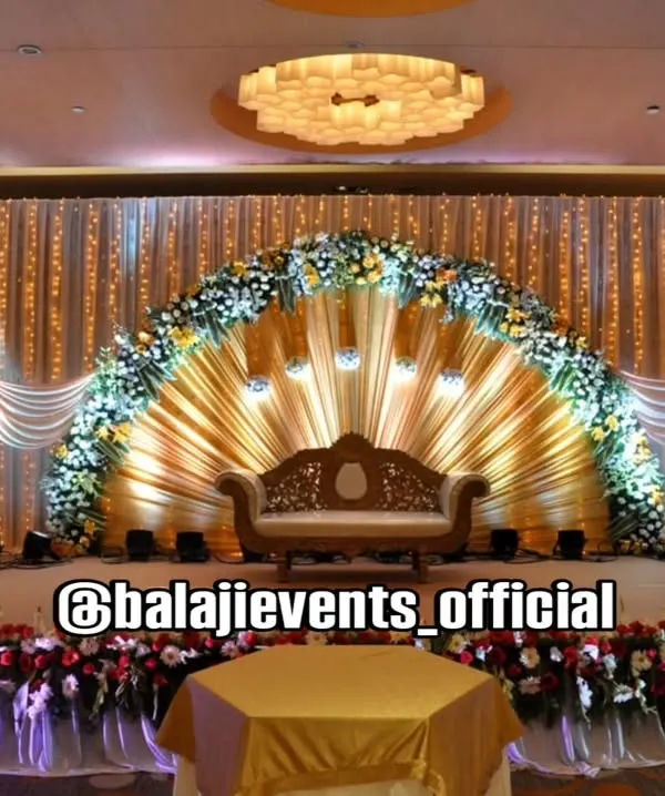 A magnificent reception stage designed to be the centerpiece of your celebration. The fan-shaped golden draping, framed by a lush floral arch and illuminated by elegant lighting, creates a truly grand and memorable backdrop for the happy couple.