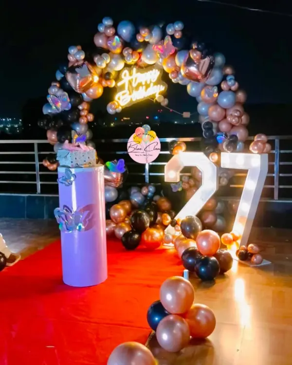A glamorous 27th birthday party setup on a rooftop, featuring a black, rose gold, and silver balloon arch with a neon sign.