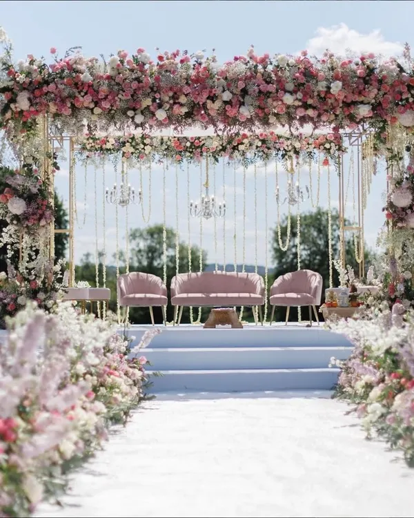 This breathtaking outdoor wedding stage is enveloped in a canopy of pink and white florals, including roses and baby's breath. The elegant pastel theme, complete with blush pink seating and delicate hanging chandeliers, creates a dreamy and romantic setting perfect for exchanging vows.