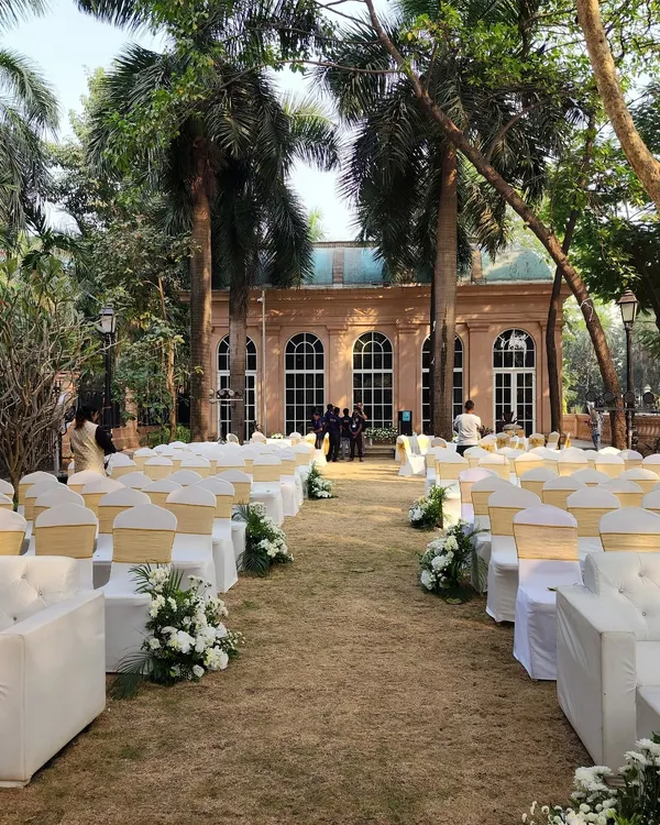 A beautiful outdoor wedding ceremony setup on a lush lawn. The aisle is lined with elegant white guest chairs and floral arrangements, leading towards a classic heritage building that serves as a stunning backdrop.