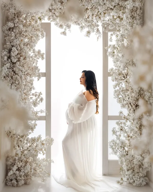 This is the single most important photo to capture your pregnancy glow. Here, a stunning mommy-to-be stands in a flowing white gown, framed by a beautiful arch of white blossoms, creating a timeless and ethereal maternity portrait.