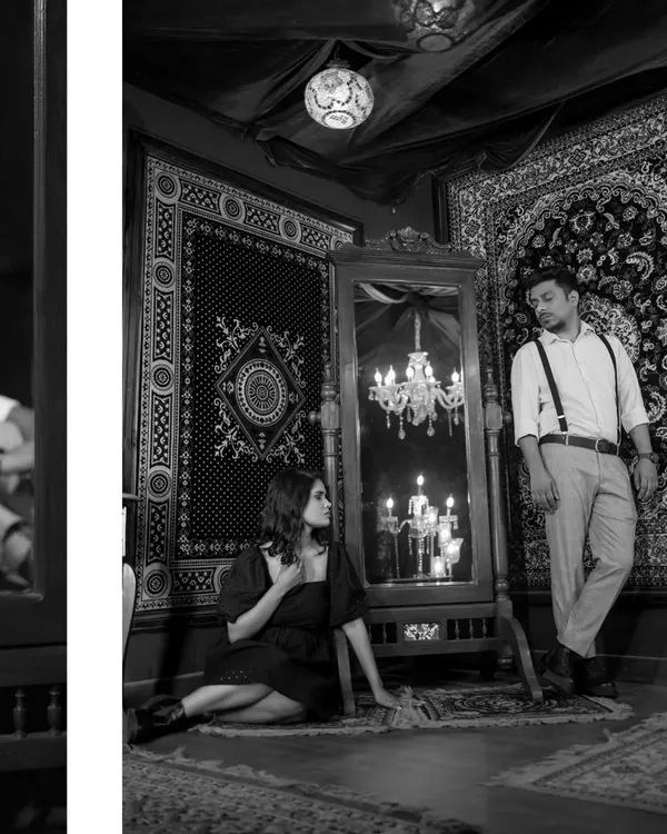 A moody black and white portrait from a stylized indoor shoot. The intricate set design, with carpets and a chandelier reflection, creates a rich, vintage, and dramatic atmosphere.