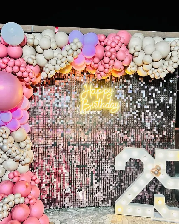 We designed this glamorous photo backdrop for a 25th birthday celebration. The setup combines a sparkling rose gold shimmer wall with a custom 'Happy Birthday' neon sign and large, light-up marquee numbers to create a perfect spot for party pictures. The scene is framed with an organic balloon arch in shades of pink and beige.