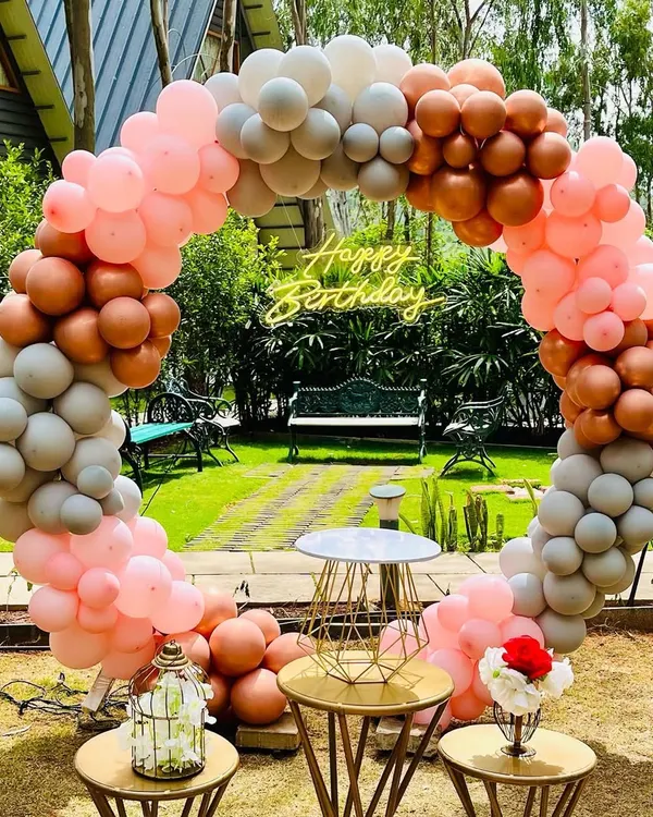 An elegant circular balloon arch in pastel pink, grey, and rose gold, set up outdoors in a garden for a beautiful birthday photo backdrop.