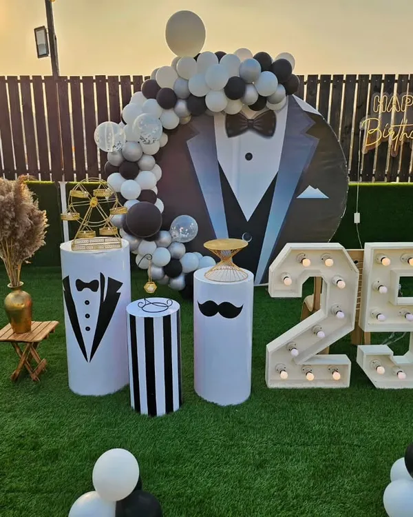 A stylish and fun "tuxedo" theme for a 25th birthday party. The decor includes a custom backdrop, themed pedestals, a balloon arch, and large light-up marquee numbers, all in a classic black and white color scheme.