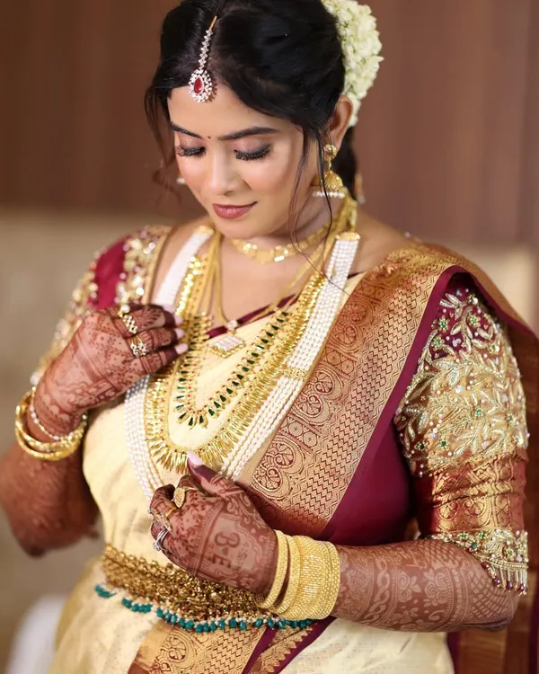 A classic South Indian bridal portrait. The focus is on her intricate gold jewelry and the rich texture of her cream and maroon Kanjeevaram saree, complemented by a soft glam makeup look.