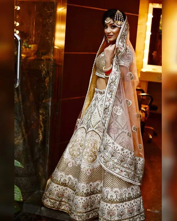 A full-length profile view of the ivory bridal lehenga, showing how the sheer pink dupatta drapes over the shoulder. The maang tikka and bridal jewelry complete the stunning look.