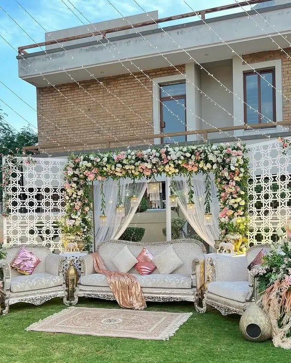 A beautiful and elegant outdoor stage setup for a daytime event. The design features a classic sofa set on a lush green lawn, framed by a floral arch in pastel pinks and whites, with intricate white lattice panels on either side.