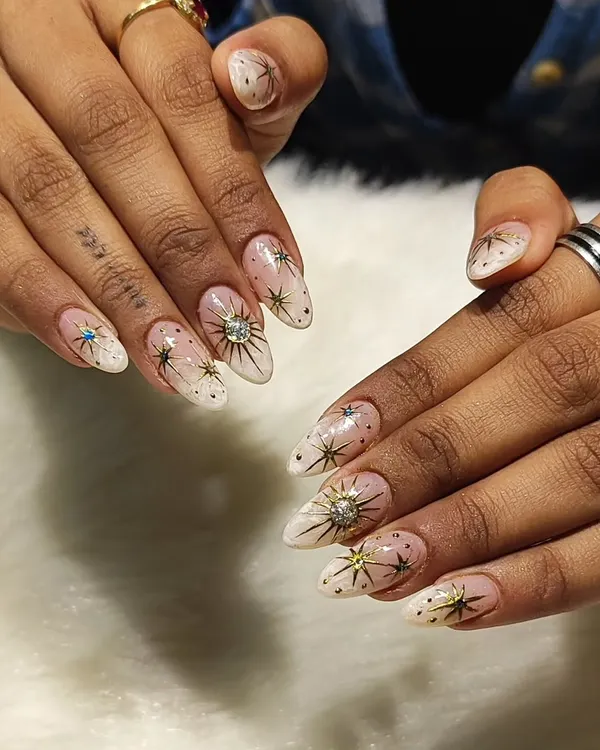 A celestial dream on your fingertips. This stunning set features a soft white ombre base on almond nails, decorated with intricate golden starbursts, tiny gold dots, and sparkling rhinestone centers.
