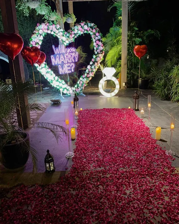 A breathtaking outdoor proposal we designed. This setup features a dense path of red rose petals leading to a large, flower-adorned heart with a "Will You Marry Me?" neon sign, all illuminated by romantic candlelight for an unforgettable moment.