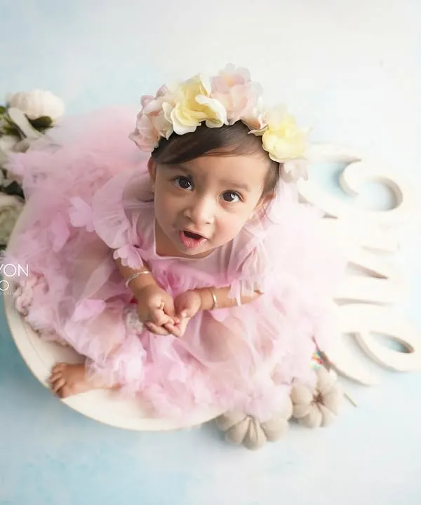 An adorable one-year-old girl in a pink tutu and floral crown, sitting in a white bowl for her first birthday portrait. The "One" sign and soft blue backdrop complete the dreamy look.